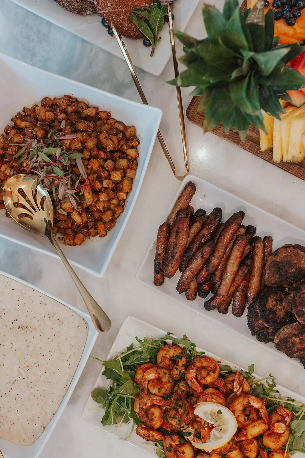 Overhead HumbledHandz brunch spread: rosemary potatoes, cheese grits, sausage, blackened shrimp with lemon, and a fruit display