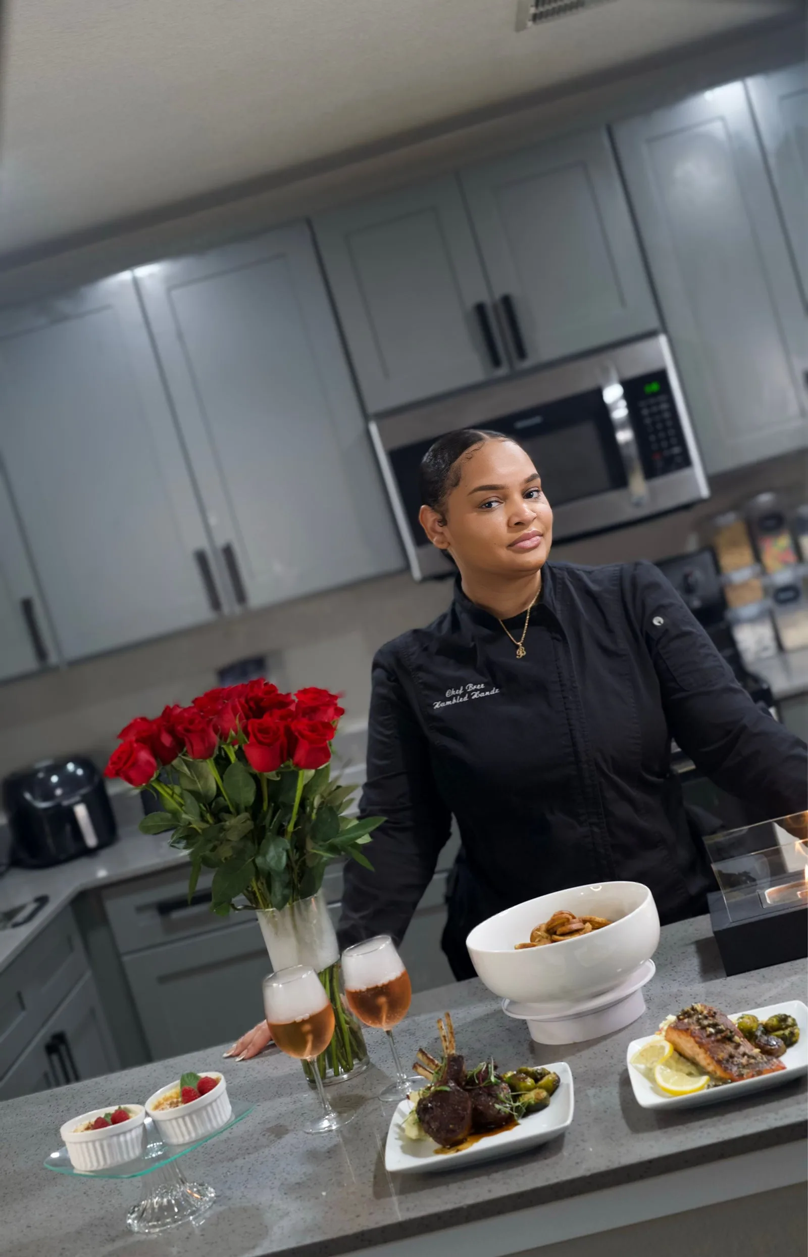 Chef Bree in her black embroidered chef coat behind plated lamb chops, salmon and creme brulee