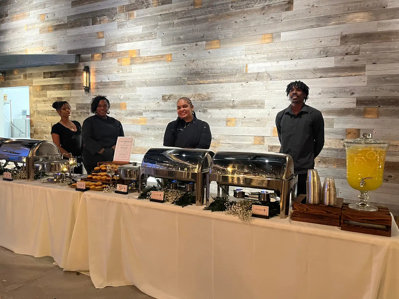 The HumbledHandz service team of four in black behind a dressed buffet line with chafing dishes and a lemonade dispenser